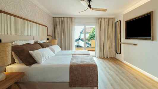 Ett hotellrum med två sängar som täcks av vita och beige sängkläder. Det finns en takfläkt, ett stort fönster med utsikt och en väggmonterad TV. (AI-genererad text)