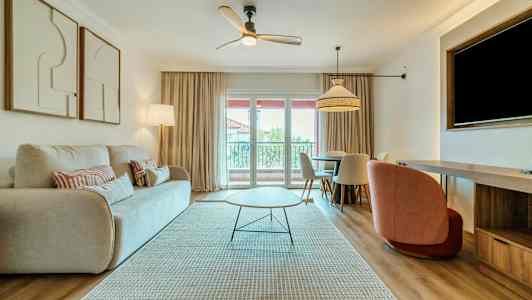 Ett vardagsrum med en ljus soffa, ett modernt soffbord, och ett matbord med stolar. Ett fönster med gardiner släpper in dagsljus. (AI-genererad text)