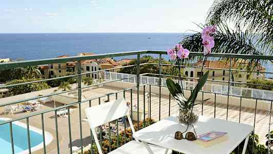 Utsikt från en balkong med bord och stolar, samt en orkidé i en kruka. En pool och byggnader i bakgrunden mot havet. (AI-genererad text)