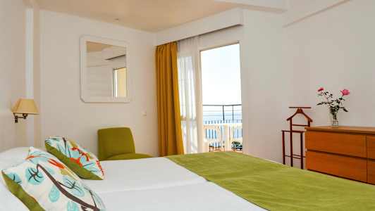 Ett hotellrum med två sängar, gröna detaljer och utsikt mot en balkong med havet i bakgrunden. (AI-genererad text)