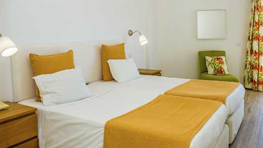 Ett hotellrum med två enkelsängar och gul sängklädsel. Ett grönt fåtölj med blommigt kudde och gardiner syns i bakgrunden. (AI-genererad text)