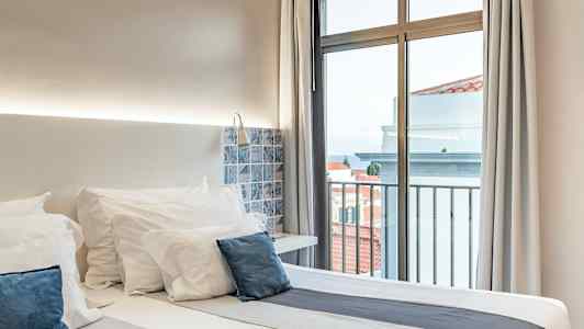 Ett hotellrum med en dubbelsäng, vita sängkläder och blå kuddar, bredvid ett fönster med utsikt. (AI-genererad text)