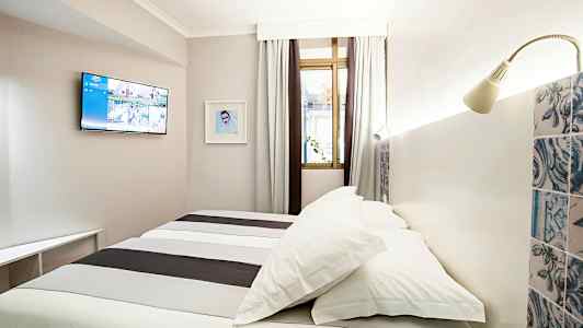 Ett hotellrum med en säng, väggmonterad TV och ett fönster med gardiner. (AI-genererad text)