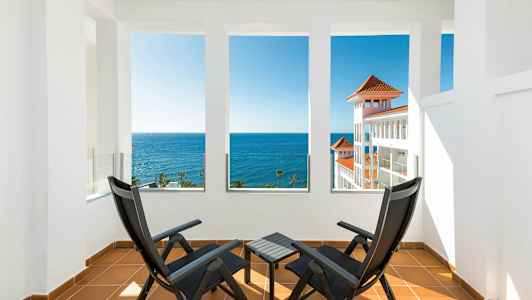 Två svarta stolar och ett bord på en terrass med utsikt över havet och en vit byggnad med röda tak. (AI-genererad text)