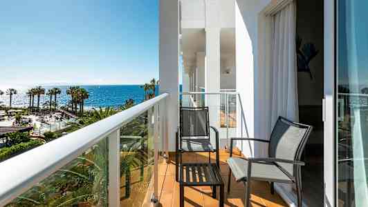 En balkong med två stolar och ett bord, med utsikt över palmer och havet. (AI-genererad text)