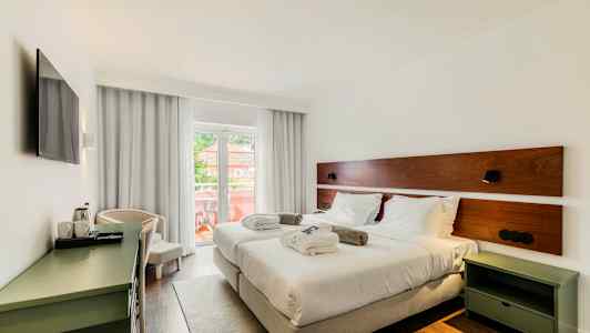 Ett hotellrum med två enkelsängar, vita sängkläder och kuddar. Rummet har en balkong med utsikt och är inrett med ett skrivbord och en TV. (AI-genererad text)