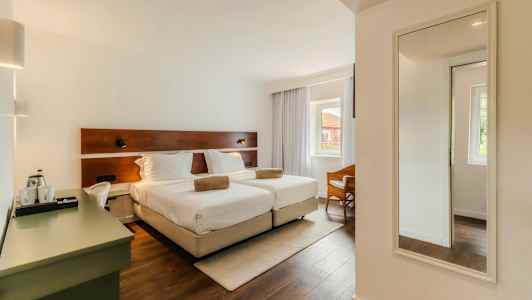 Ett hotellrum med två enkelsängar, en skrivbordshörna och en spegel på väggen. Rummet har ett stort fönster med gardiner. (AI-genererad text)