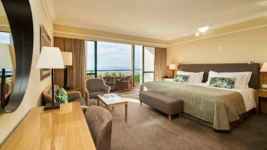 Ett hotellrum med säng, sittgrupp och utsikt mot balkong och hav. (AI-genererad text)