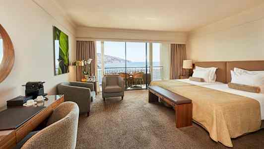 Hotellrum med dubbelsäng, sittgrupp och balkong med utsikt över havet. (AI-genererad text)