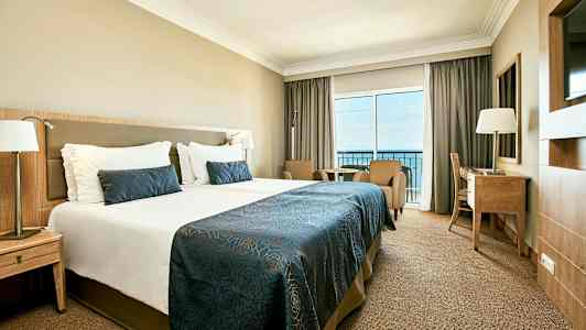 Bild av ett hotellrum med en stor säng, två fåtöljer, ett skrivbord och en balkong som visar utsikt över havet. (AI-genererad text)