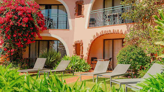 En byggnad med välvda balkonger omgiven av grönska och blommor, med solstolar placerade på gräsmattan. (AI-genererad text)
