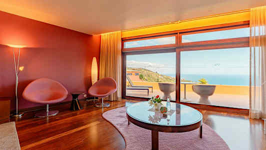 Ett rum med röda väggar, två stolar, ett bord och stora fönster som vetter mot en terrass med havsutsikt. (AI-genererad text)