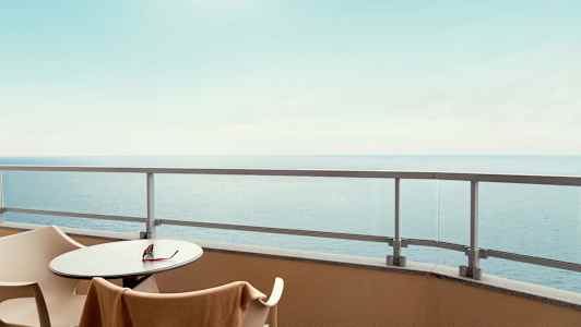 Utsikt från en balkong med bord, stolar och glasräcke, havet syns i bakgrunden. (AI-genererad text)
