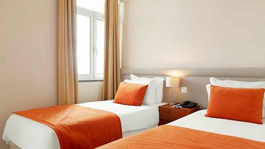 Ett hotellrum med två enkelsängar bredvid varandra, med vita lakan och orange överkast och prydnadskuddar. (AI-genererad text)