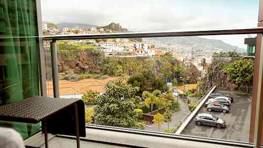 Utsikt från en balkong med möbler mot en bergig miljö med byggnader och parkerade bilar. (AI-genererad text)