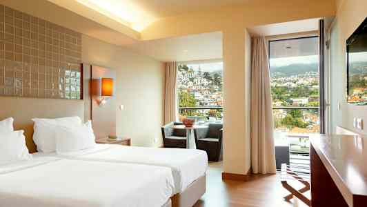 Ett hotellrum med två enkelsängar, en sittgrupp vid ett fönster med utsikt över en stad i en bergig miljö. (AI-genererad text)