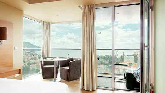 Hotellrum med panoramavy genom fönster och dörrar över en stad framför havet. (AI-genererad text)
