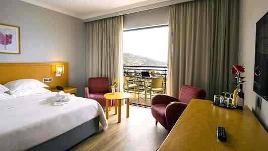 Hotellrum med en dubbelsäng, två röda fåtöljer, ett skrivbord med drycker och glas, och en balkong med utsikt. (AI-genererad text)