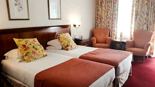 Ett hotellrum med två enkelsängar, dekorerade med blommiga kuddar och röda överkast. I rummet finns också två fåtöljer, ett litet bord och gardiner med blommönster. (AI-genererad text)