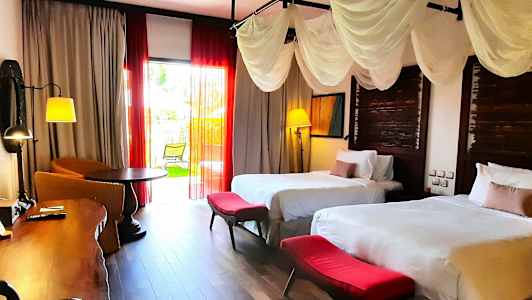 Ett hotellrum med två sängar, vita sängkläder och röda fotpallar. Rummet har dekorativa gardiner, ett skrivbord, en sittgrupp och en balkongdörr som leder ut till en grönskande trädgård. (AI-genererad text)