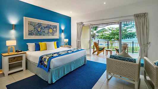 Ett hotellrum med en stor säng, blå och gula dekorativa kuddar, en balkong med utsikt över havet och omgivande grönska. (AI-genererad text)