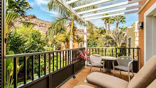 En balkong med två solstolar, bord och stolar. Utsikt mot grönskande trädgård och berg. (AI-genererad text)