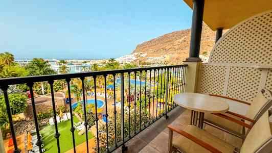 Utsikt från balkong över poolområde med grönska, palmer och en bergskulle i bakgrunden. (AI-genererad text)