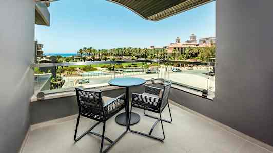 Balkong med två stolar och ett bord, utsikt mot grönt område och hav. (AI-genererad text)