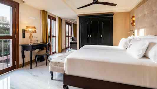 Ett hotellrum med dubbelsäng, svart garderob, guldfärgad bordslampa och fönster med gulddraperier. (AI-genererad text)