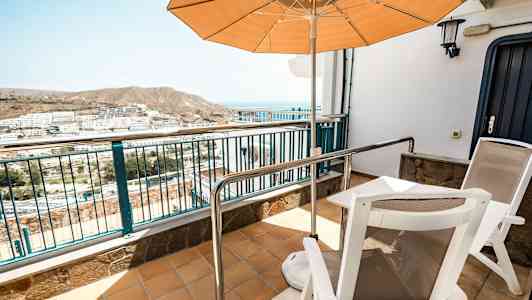 En terrass med två stolar, ett bord, och ett parasoll, med utsikt över ett bostadsområde och berg i bakgrunden. (AI-genererad text)