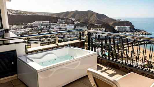 En terrass med bubbelbad och utsikt över en bukt med hotell och strand. (AI-genererad text)