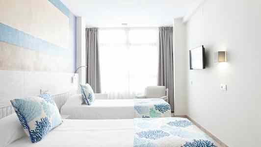 Två enkelsängar med blå och vita sängkläder i ett ljust rum med ett stort fönster. (AI-genererad text)