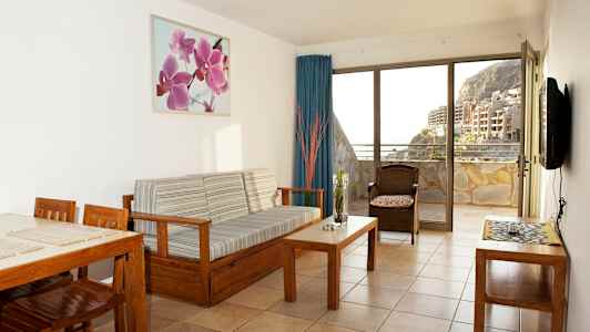 Ett vardagsrum med träinredning och utsikt över en balkong samt byggnader och berg. (AI-genererad text)