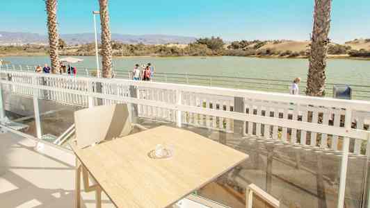 Utsikt från en terrass med bord och stol, med vatten och gångväg i bakgrunden. (AI-genererad text)