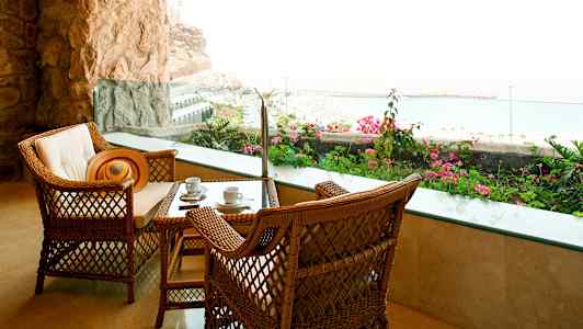 Två rottingstolar och ett bord med kaffekoppar, på en terrass med utsikt över havet och blomsterprakt. (AI-genererad text)