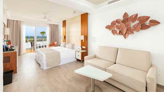 Ljust hotellrum med säng, soffa och balkong samt havsutsikt. (AI-genererad text)