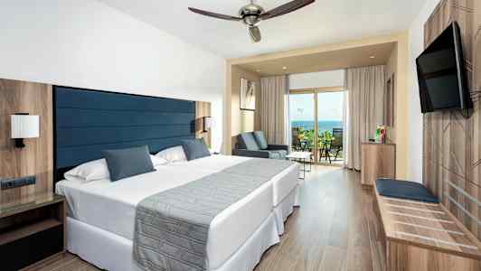 Ett hotellrum med två sängar, ett sittområde och en balkong som har utsikt över havet. (AI-genererad text)