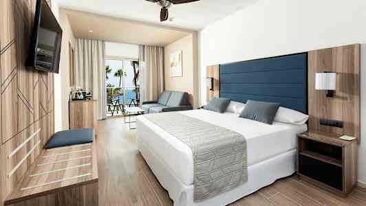 Ett hotellrum med en stor säng, en soffa och ett bord. Det finns en balkong med utsikt över havet. (AI-genererad text)