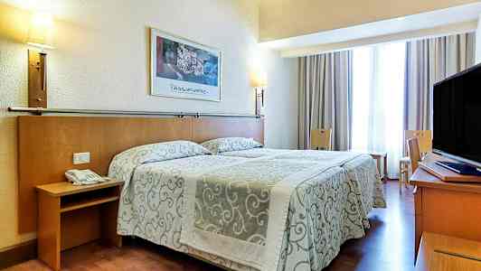 Ett hotellrum med en dubbelsäng med mönstrade överkast, lampor på väggen och en TV på ett skrivbord. Sittmöbler och ett fönster med gardiner finns också. (AI-genererad text)