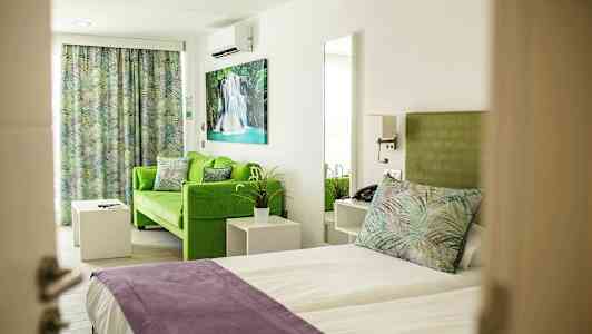 Rum med säng och grön soffa, med färgglada kuddar och gardiner samt konst på väggen. (AI-genererad text)