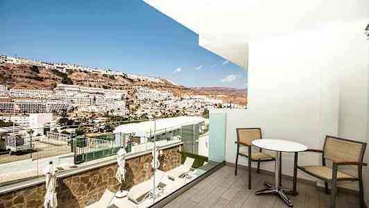 Utsikt från en balkong med bord och stolar ut mot en sluttning med byggnader i fjärran. (AI-genererad text)