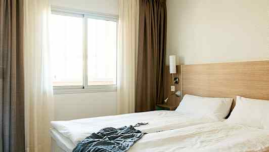 Ett hotellrum med en dubbelsäng, vita sängkläder, en trävägg vid sänggaveln och ett fönster med ljusa gardiner. (AI-genererad text)