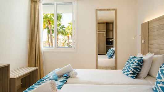 Ett hotellrum med två enkelsängar, blå kuddar, spegel och utsikt över palmer genom fönstret. (AI-genererad text)