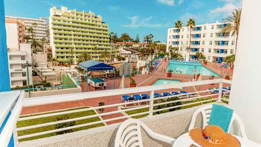 Utsikt från en balkong som visar ett hotellområde med pool och solstolar samt byggnader i bakgrunden. (AI-genererad text)