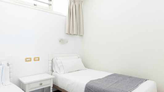 Två enkelsängar med vita sängkläder och en grå filt, belägna i ett ljust rum. (AI-genererad text)