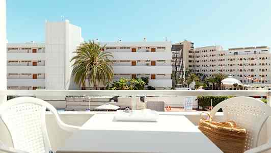 Utsikt från en balkong med sittgrupp, byggnader och palmer synliga i bakgrunden under en klarblå himmel. (AI-genererad text)