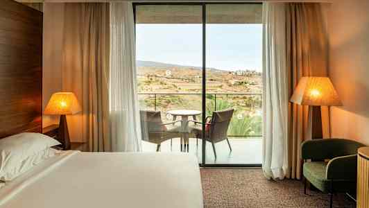 Ett hotellrum med en säng, två bordslampor, en fåtölj och en balkong med sittmöbler. (AI-genererad text)