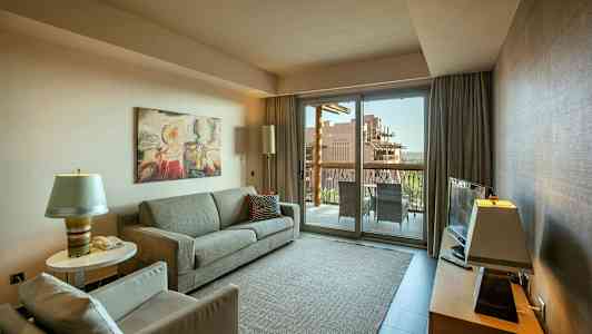 Ett vardagsrum med en soffa, fåtölj, och ett soffbord, en TV på en möbel samt en glasdörr som leder ut till en balkong. (AI-genererad text)