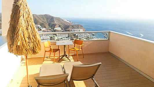 Utsikt från en terrass med parasoll, två stolar och ett bord, samt solstolar med en havsutsikt. (AI-genererad text)