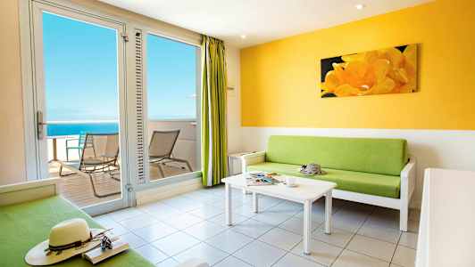 Ett ljust vardagsrum med grön soffa, vitt soffbord och utgång till balkong som har utsikt över havet. (AI-genererad text)